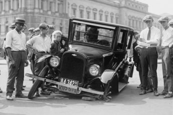 A black and white old photo of people posing beside a flat tired black car in the road