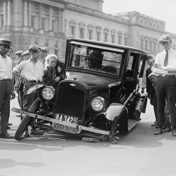 A black and white old photo of people posing beside a flat tired black car in the road