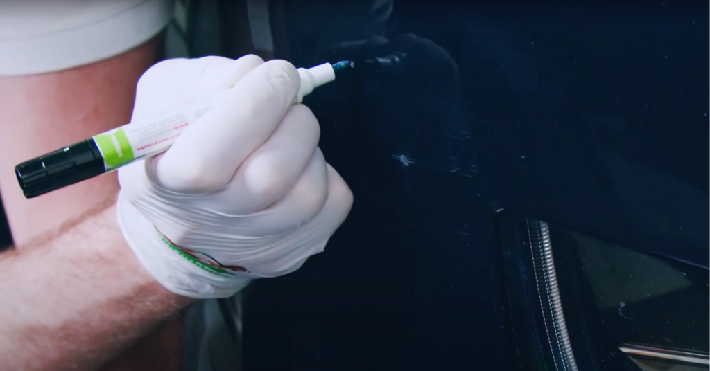 A gloved hand touches up a black car with a TouchUpDirect Touch Up Pen.