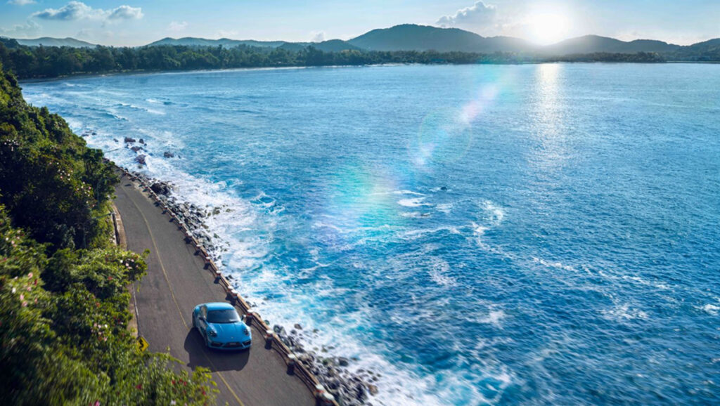 A Porsche in Riviera Blue drives on a road next to a sparkling ocean on a sunny day.