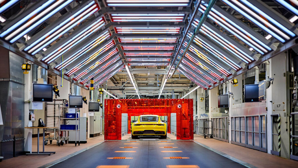 A yellow Porsche sits on the floor of the plant at Zuffenhausen, a rainbow arch of lights above it.