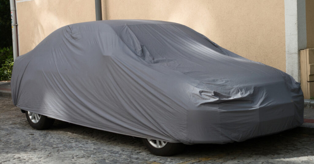 A car parked outside on a stone street is protected by a gray car cover.