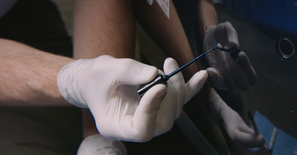 A hand delicately touches up a scratch on a black car with a touch up Brush.