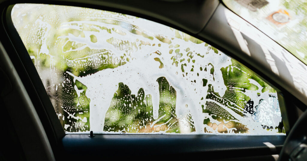 A view from the inside of a car as someone washes the outside driver's seat window with soap.