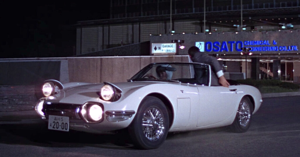 James Bond gets into small white Toyota convertible driven by Aki with the building and logo of Osato Chemical & Engineering Co., Ltd. in the background.