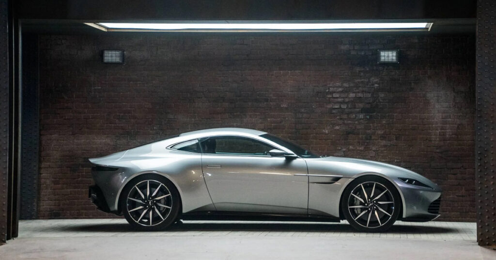 A silver Aston Martin DB10 parked in front of a brick wall, under flattering lights.